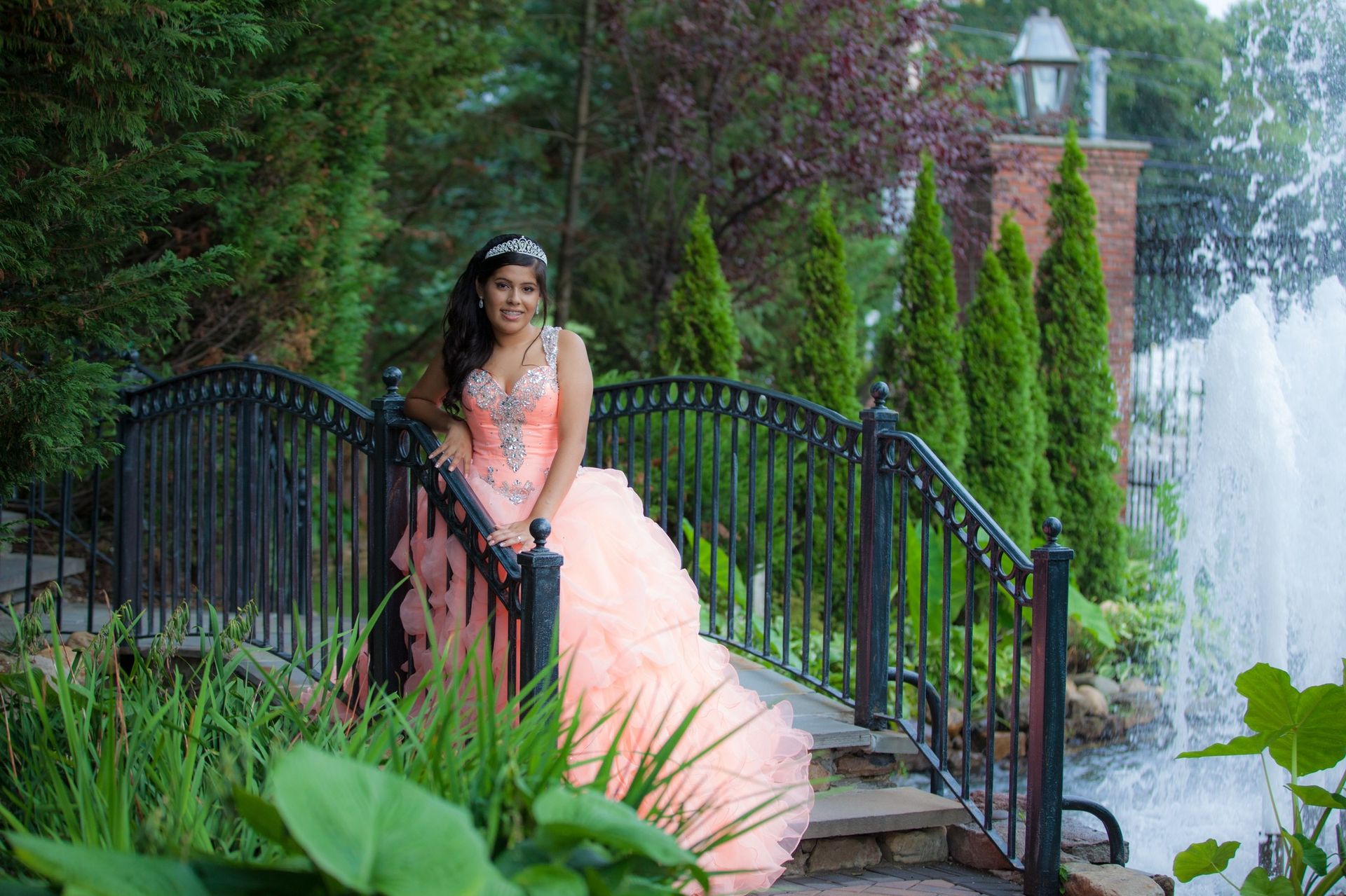 Quinceañera in a peach gown posing on a garden bridge beside a fountain