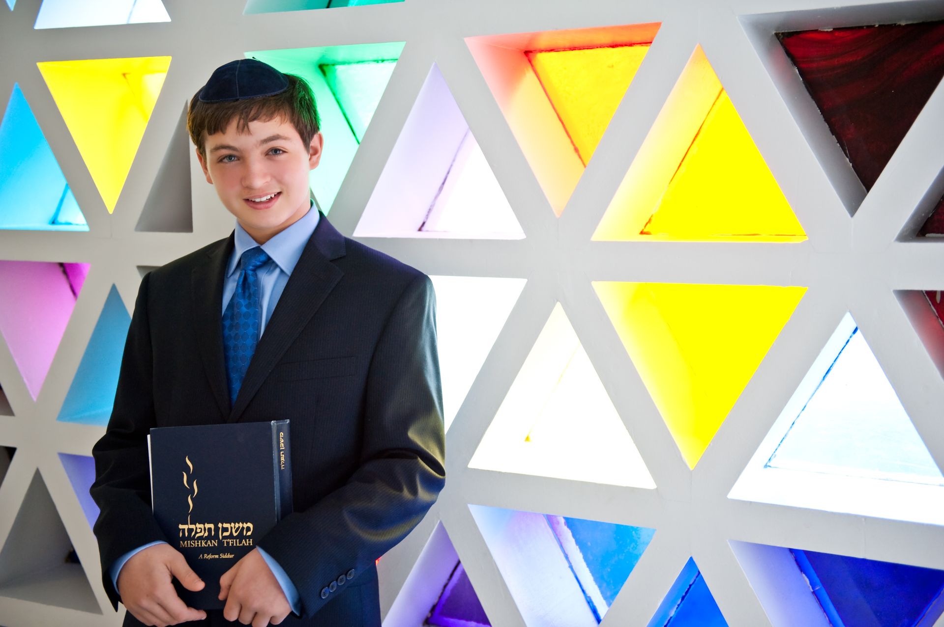 Bar mitzvah boy in a blue kippah and suit holding a siddur before a colorful geometric stained-glass wall