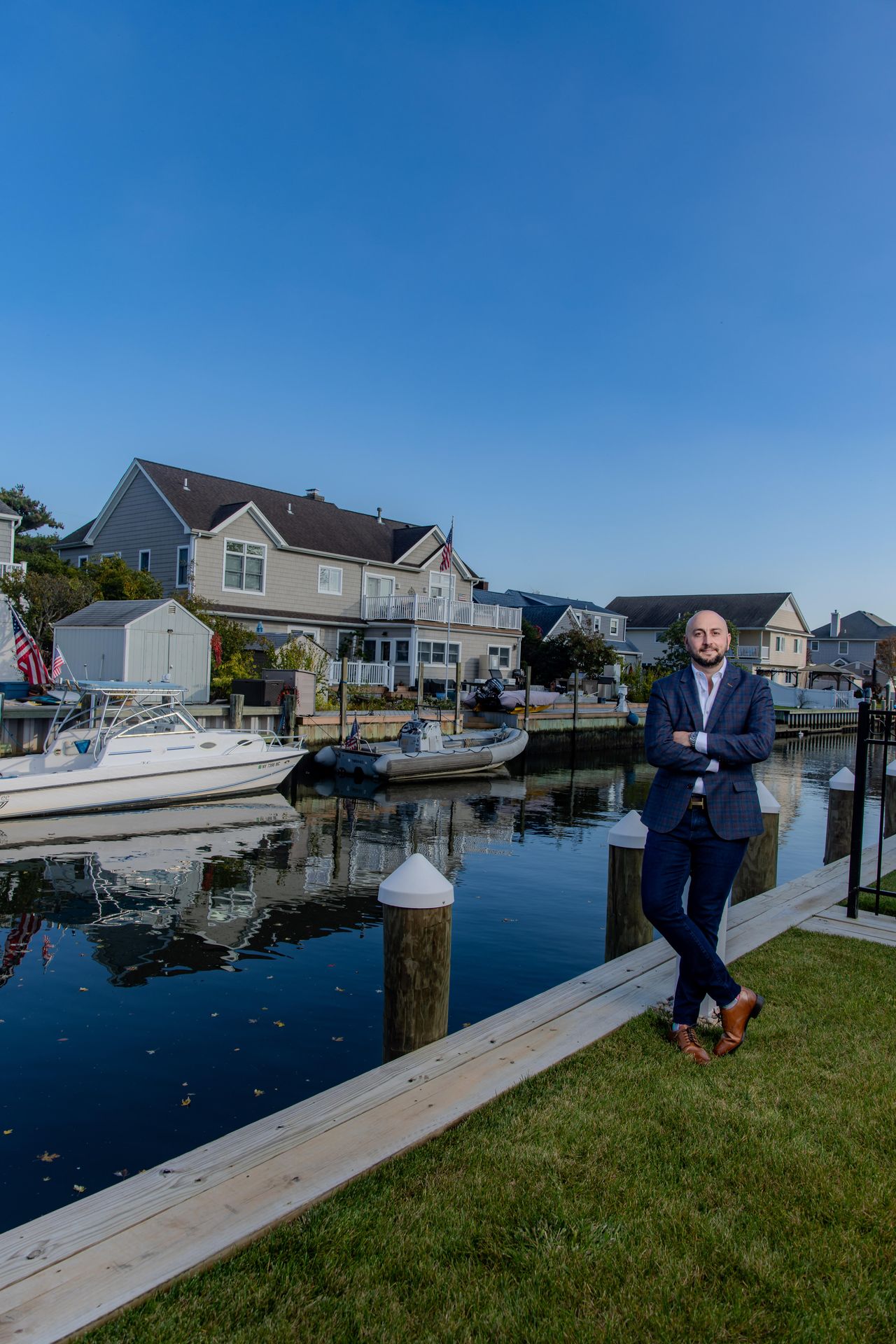 Bald man in a blue plaid blazer leaning against a dock post by the water, waterfront houses and boats behind him