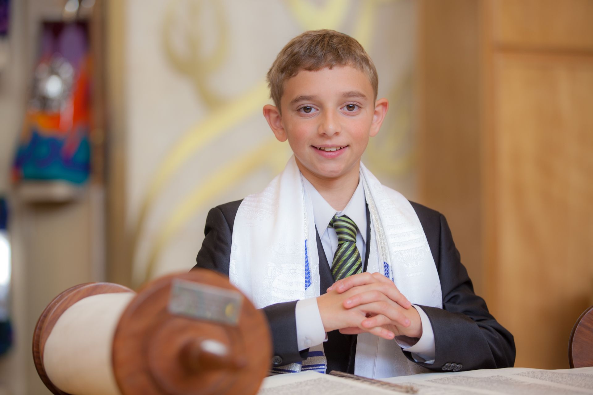 Bar mitzvah boy in a green tie and tallit seated at a table with a Torah scroll beside him