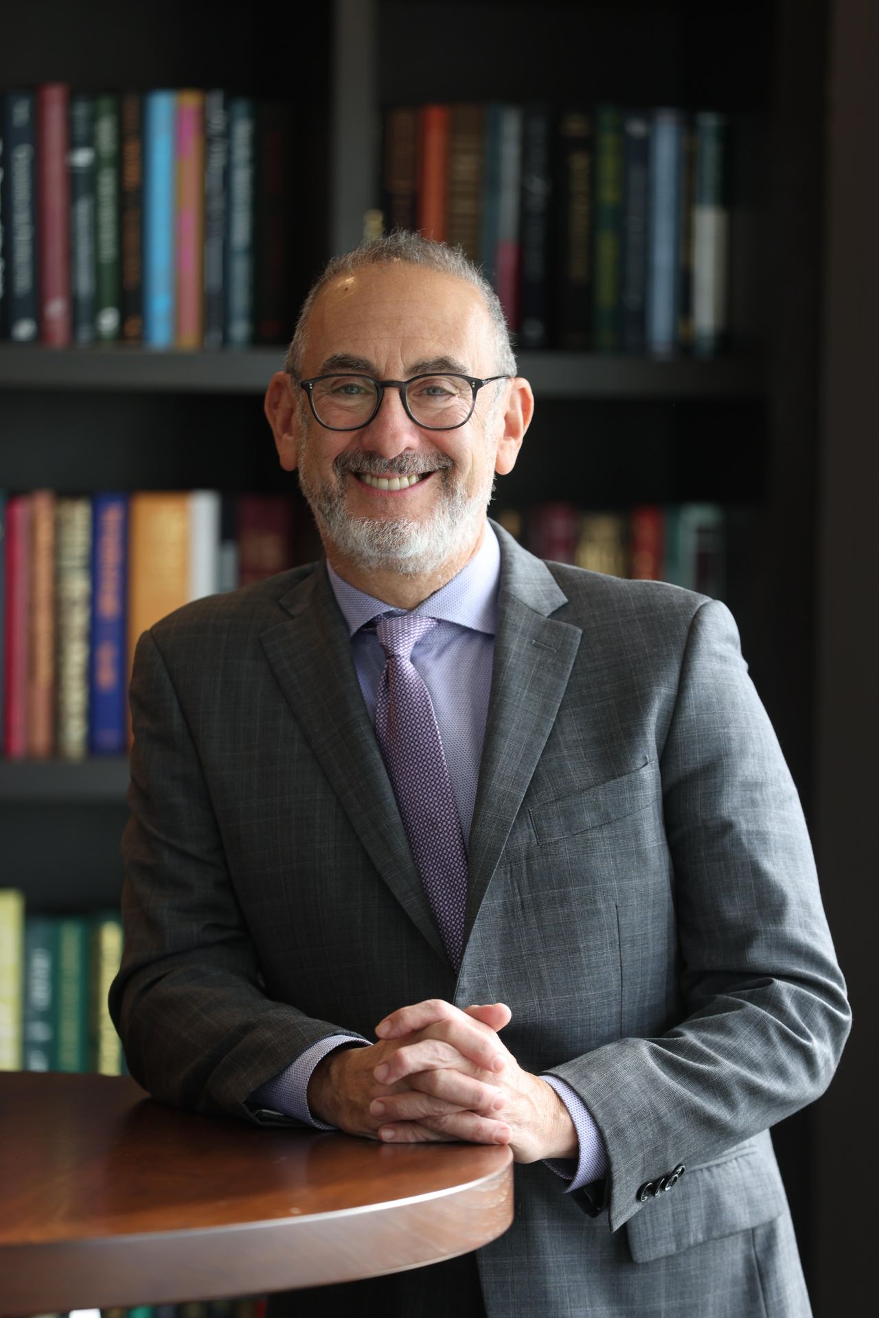 Smiling bearded man with glasses in a gray suit leaning on a desk before a bookshelf