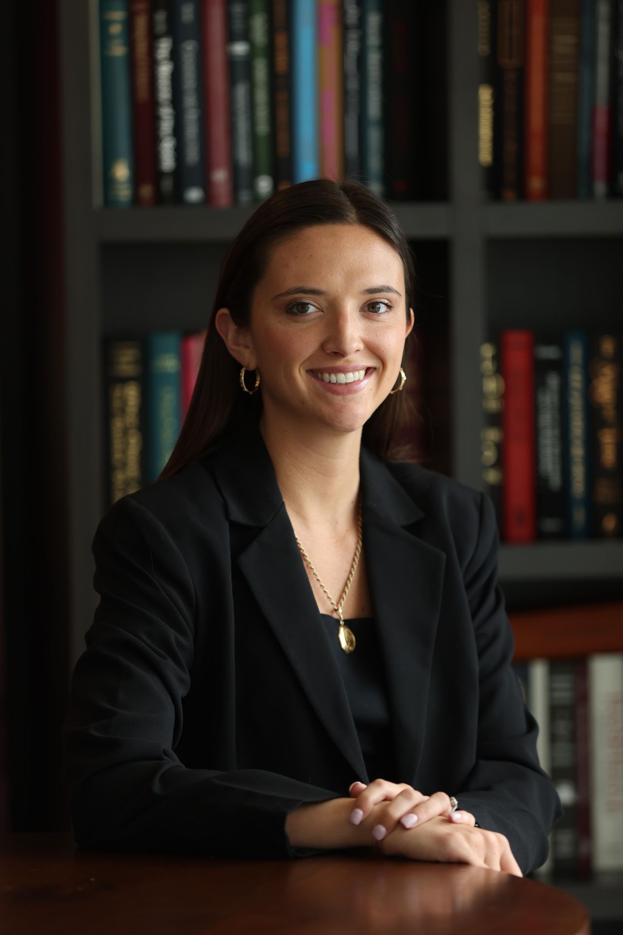 Smiling woman in a black blazer seated at a desk before a bookshelf