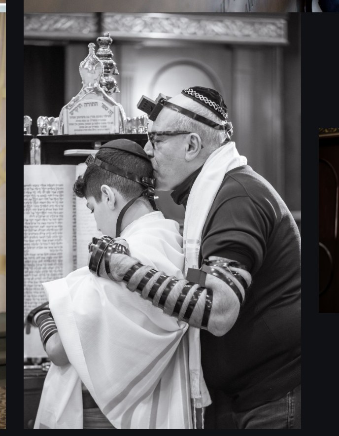 Black-and-white photo of a father kissing his bar mitzvah son's head, both wearing tefillin and tallit at the Torah